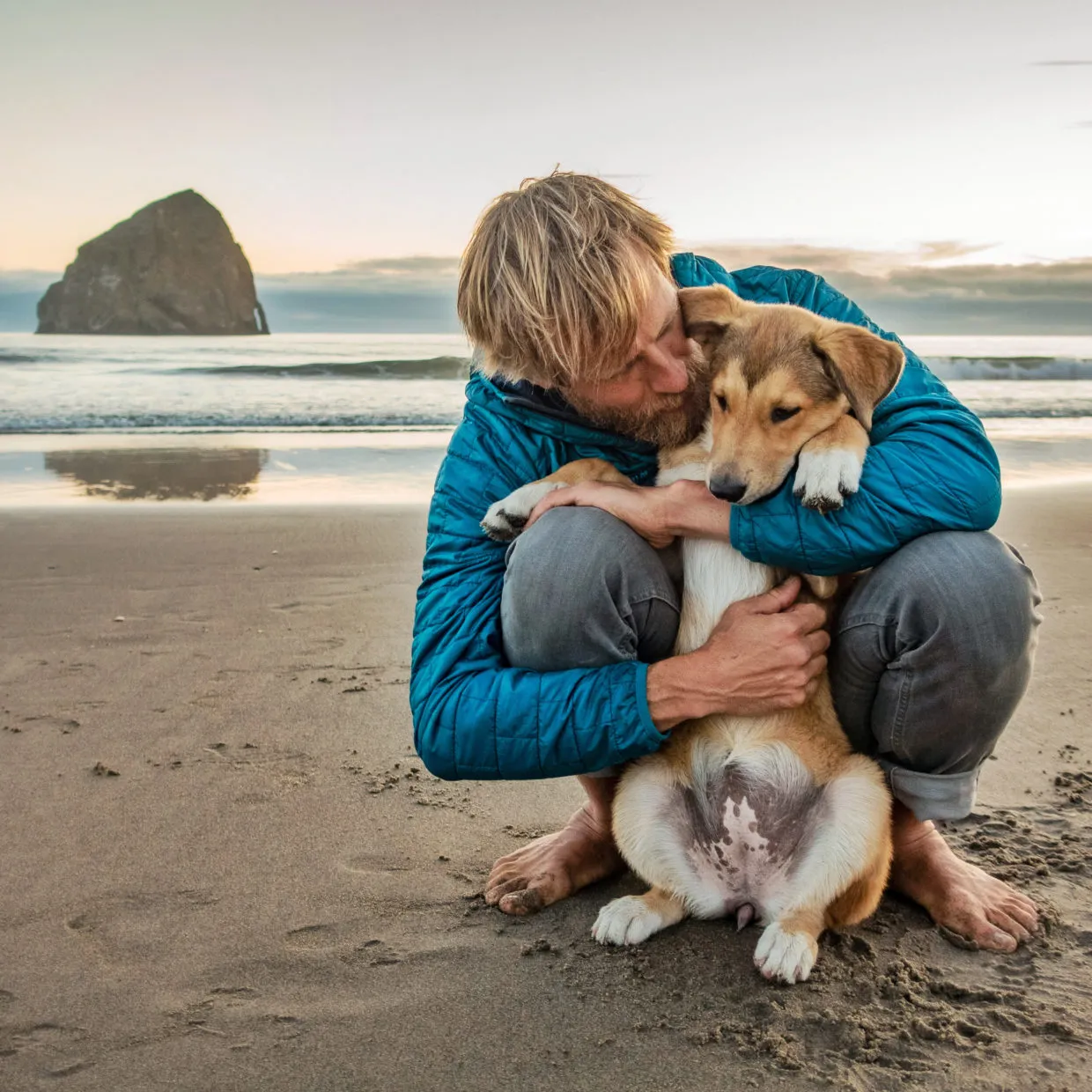 Ben Moon sits on a rocky beach, holding a fluffy puppy named Nori, with the ocean in the background