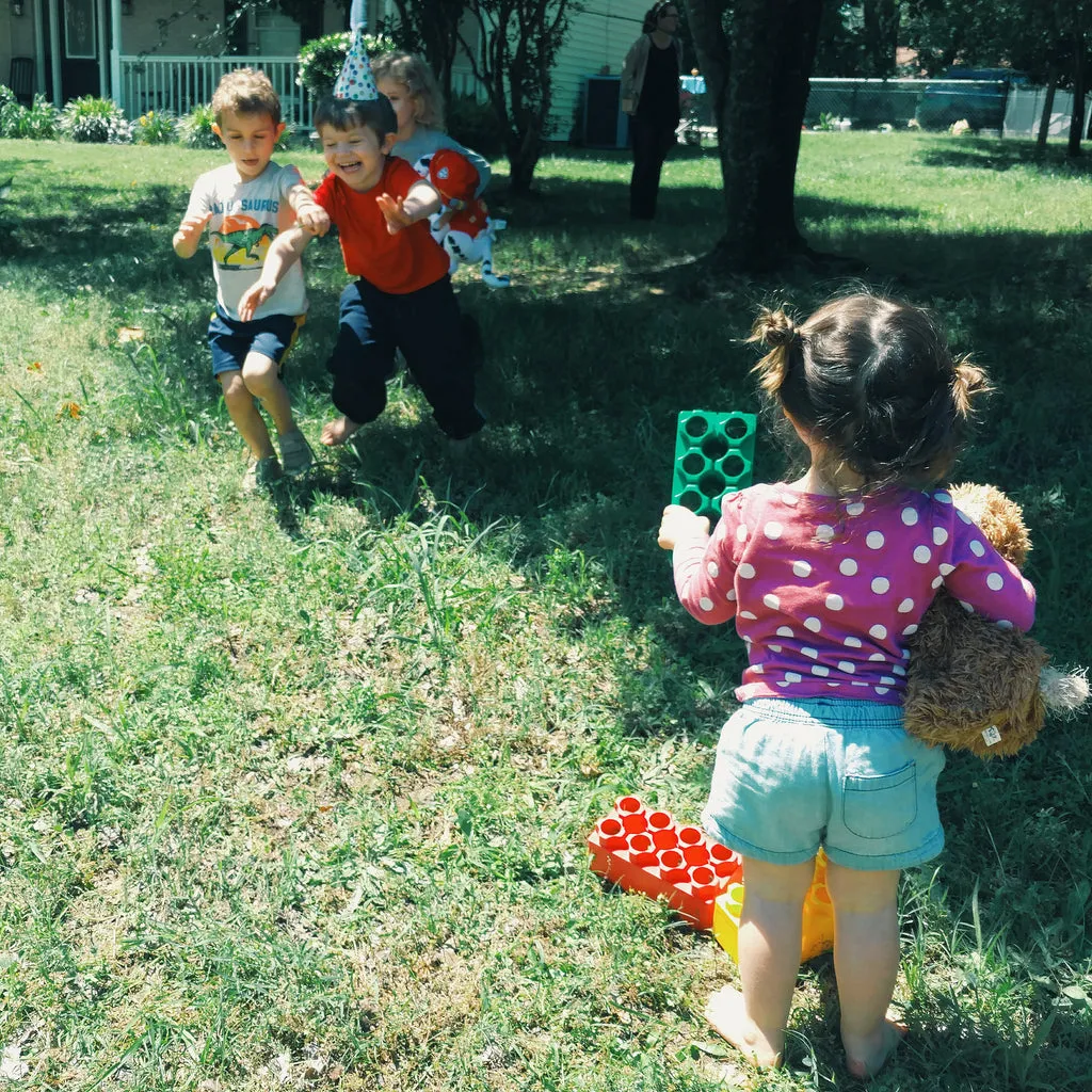 Children playing an outdoor game of "Red Light, Green Light" with an adult holding up colored blocks as visual cues, tying into the themes of the "Go, Dog. Go!" book.