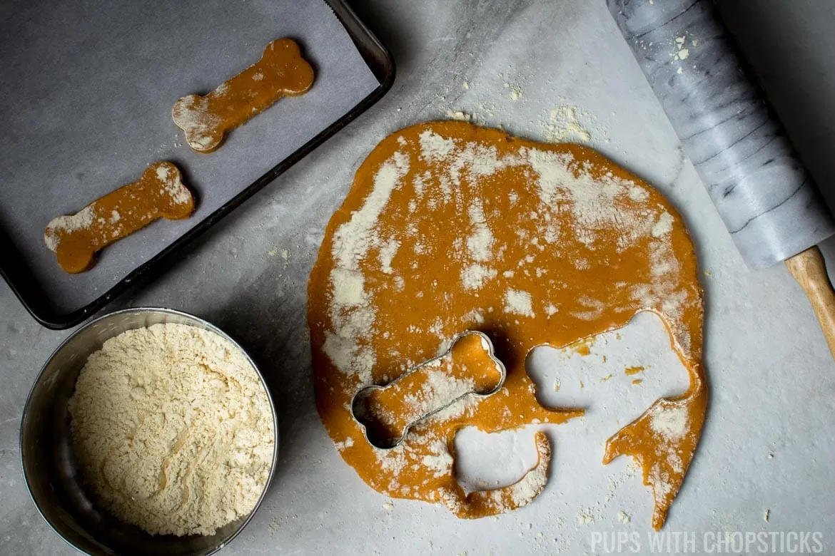 Grain-free pumpkin dog treat dough being rolled out and cut into bone shapes using a cookie cutter on a floured surface.