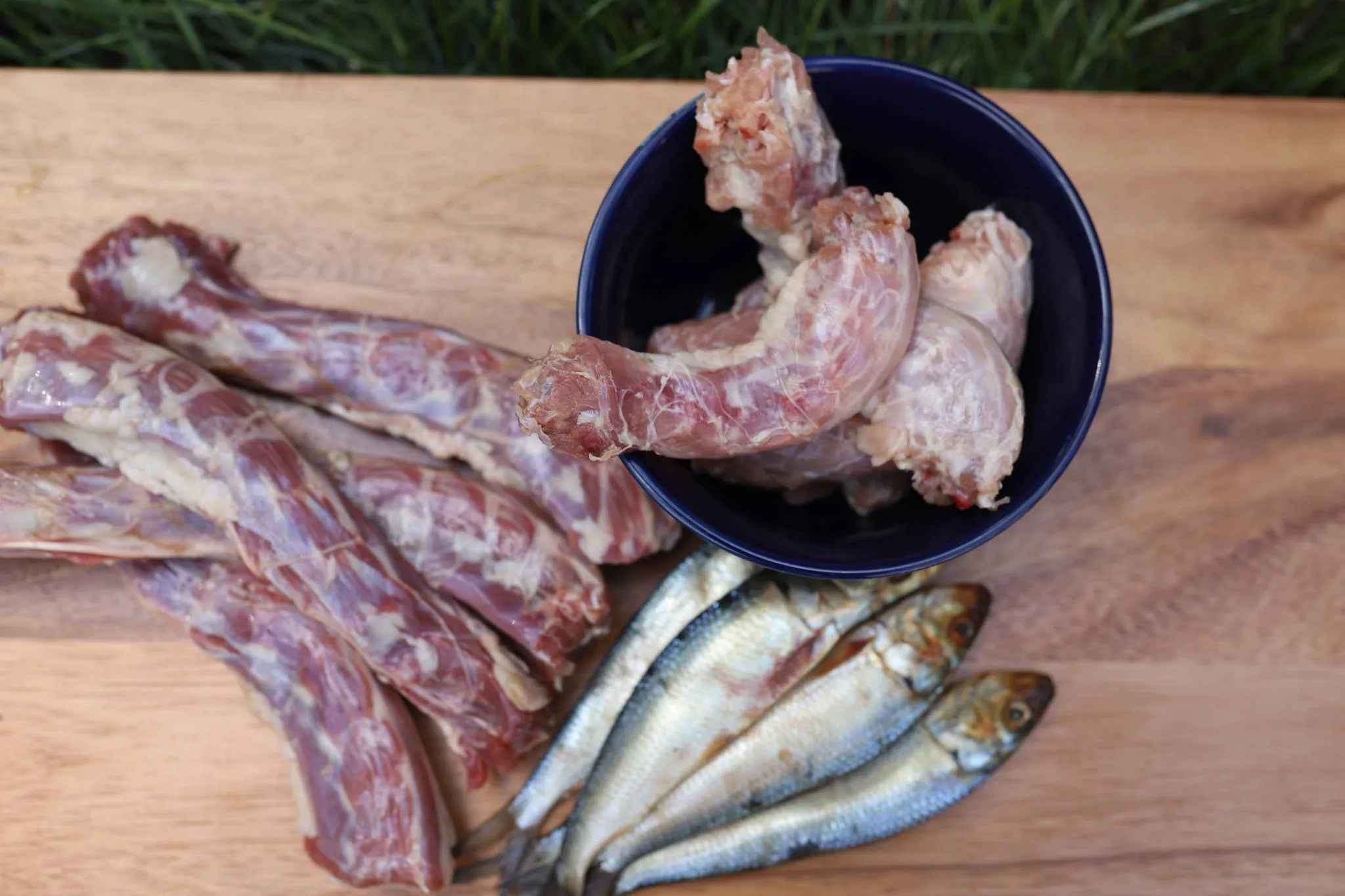 Variety of raw meaty bones (chicken necks, turkey necks, sardines) displayed on a wooden board, ideal for dog nutrition.