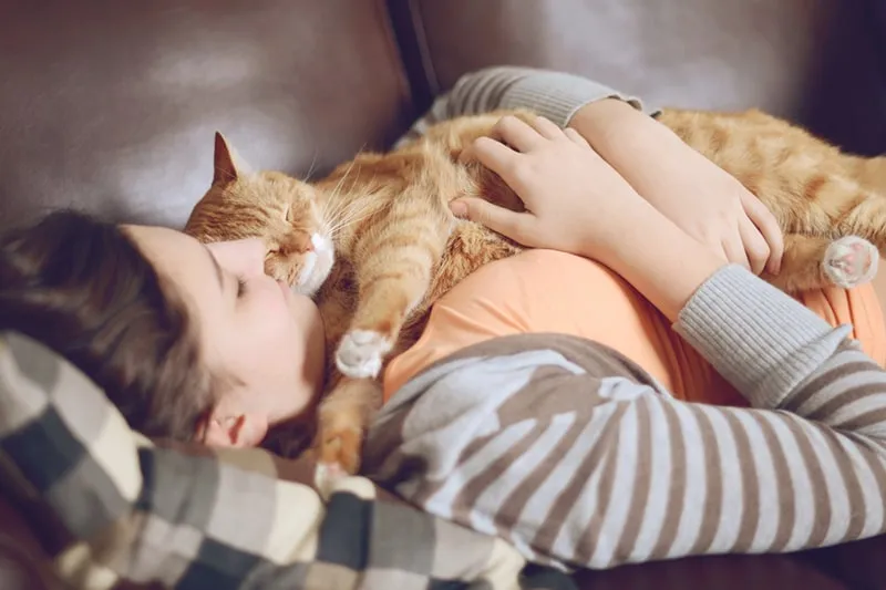 Woman lovingly cuddling an orange tabby cat on a brown leather sofa, illustrating the bond with pets and the importance of managing the average monthly cost for a cat.