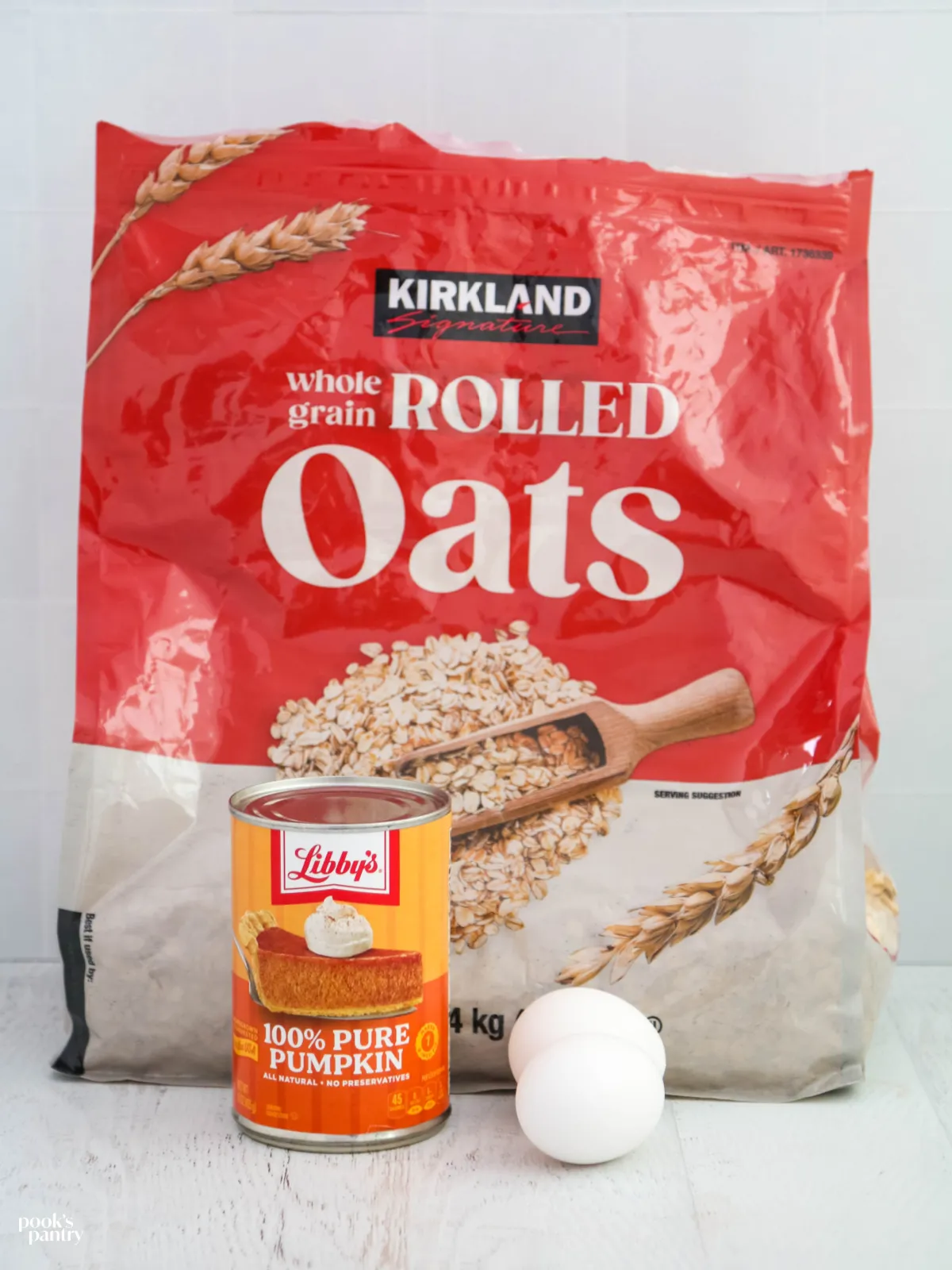 A bag of old-fashioned oats, a can of pure pumpkin purée, and two eggs are displayed on a clean white background, ready for making homemade dog treats.