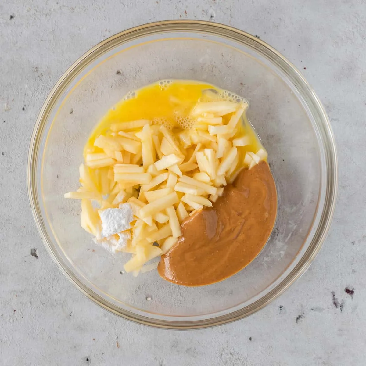 A collection of ingredients for the dog cake: chopped apple, peanut butter, a raw egg, and baking powder in a glass bowl before mixing