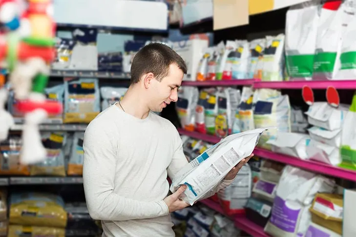 A man in a supermarket aisle carefully reads the nutritional label on a large bag of dog food.