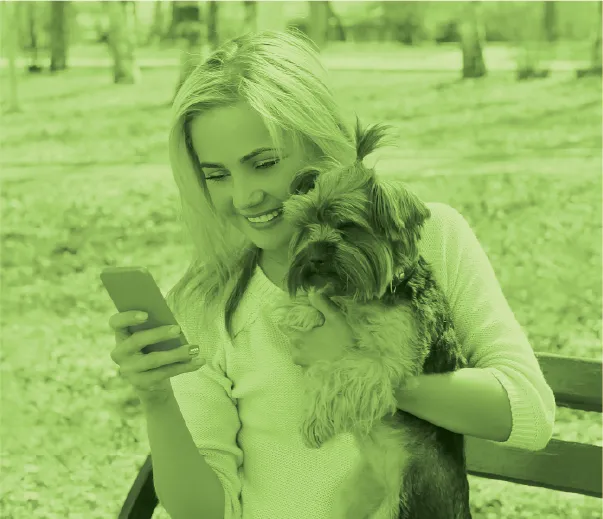 A millennial woman smiling while holding her dog and looking at her smartphone
