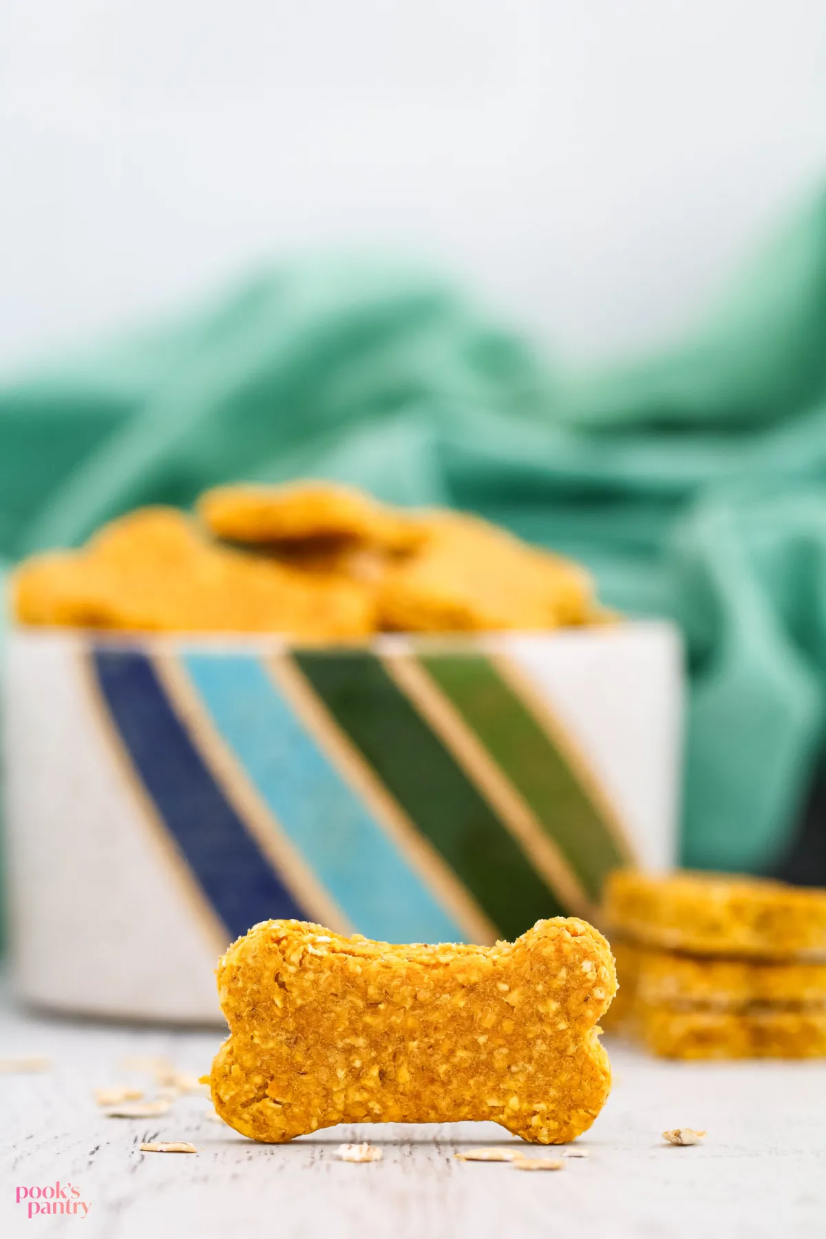 A single homemade oatmeal pumpkin dog treat stands on its side in front of a pottery bowl filled with more of the wholesome dog biscuits.