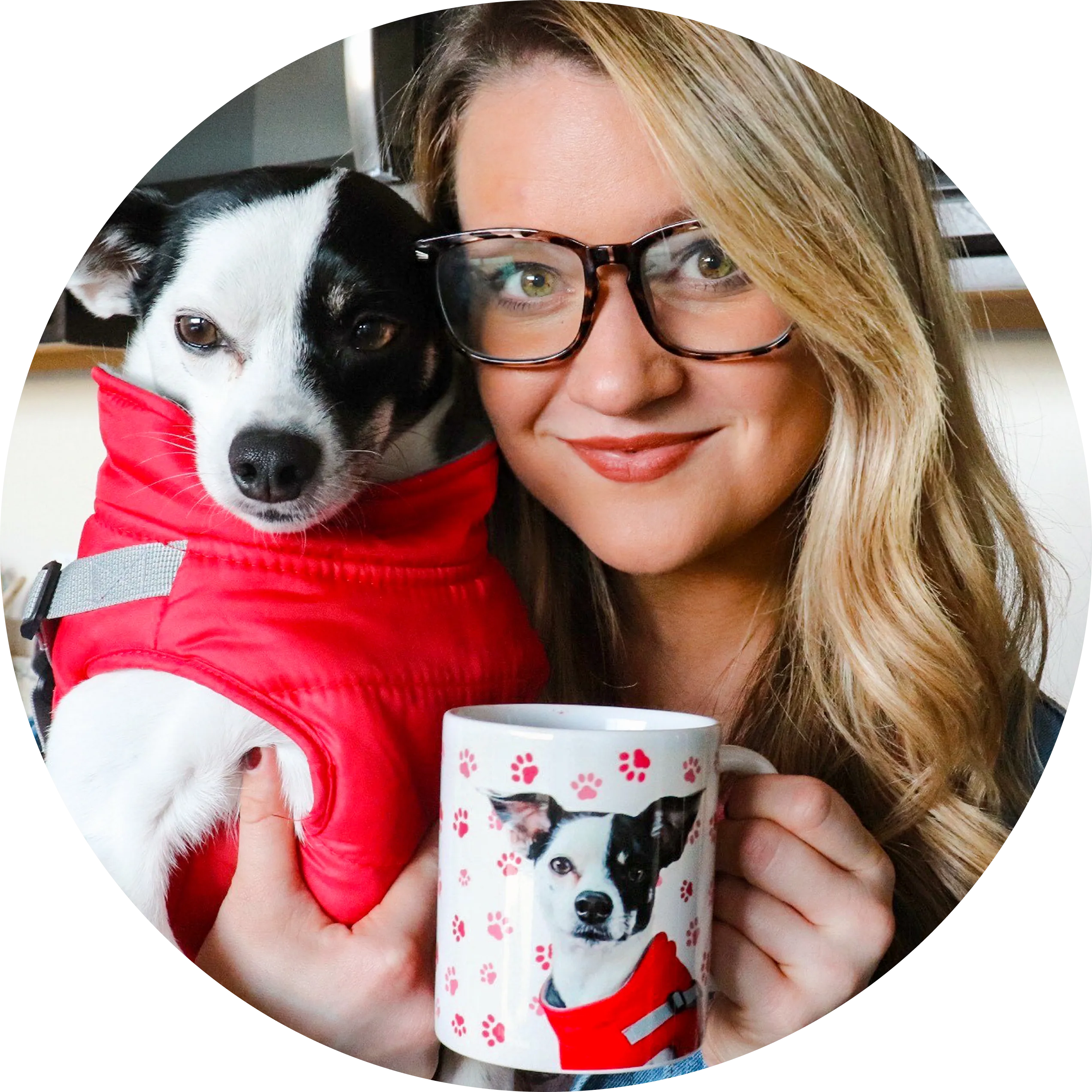 A smiling woman with blonde hair and tortoise-shell glasses is holding a black and white Terrier mix dog wearing a red puffer vest. The woman is also holding a white mug patterned with red and pink paw prints and a portrait of the dog wearing the matching red vest.