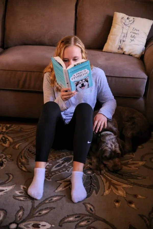 A teenager reads a book while cuddling with a dog on a couch