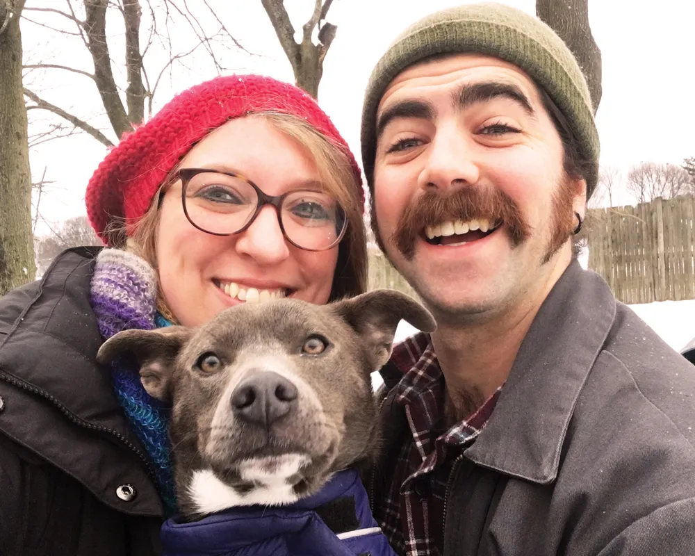 A worn green dog leash hangs on a white wall, with two dog collars, one blue and one pink with paw prints, hooked onto the same nail.