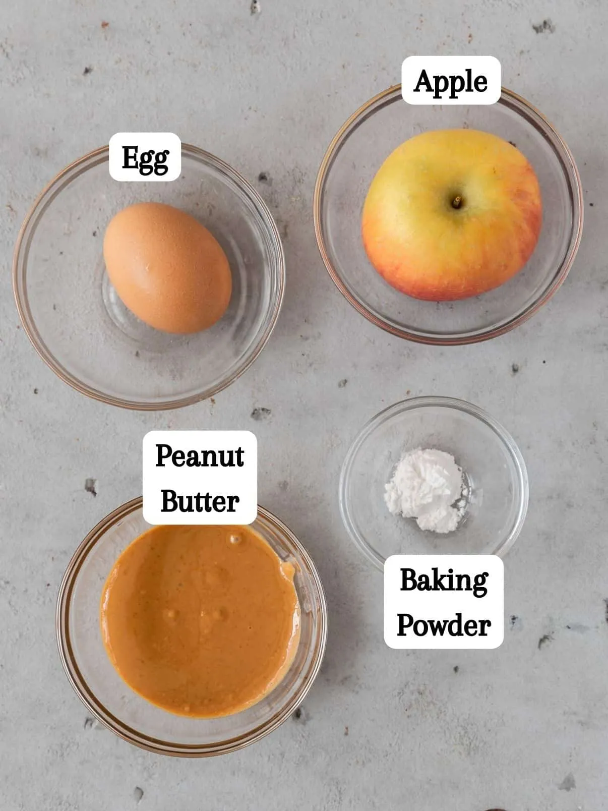 All the raw ingredients for a homemade dog cake, including an egg, peanut butter, chopped apple, and baking powder, laid out in glass bowls.
