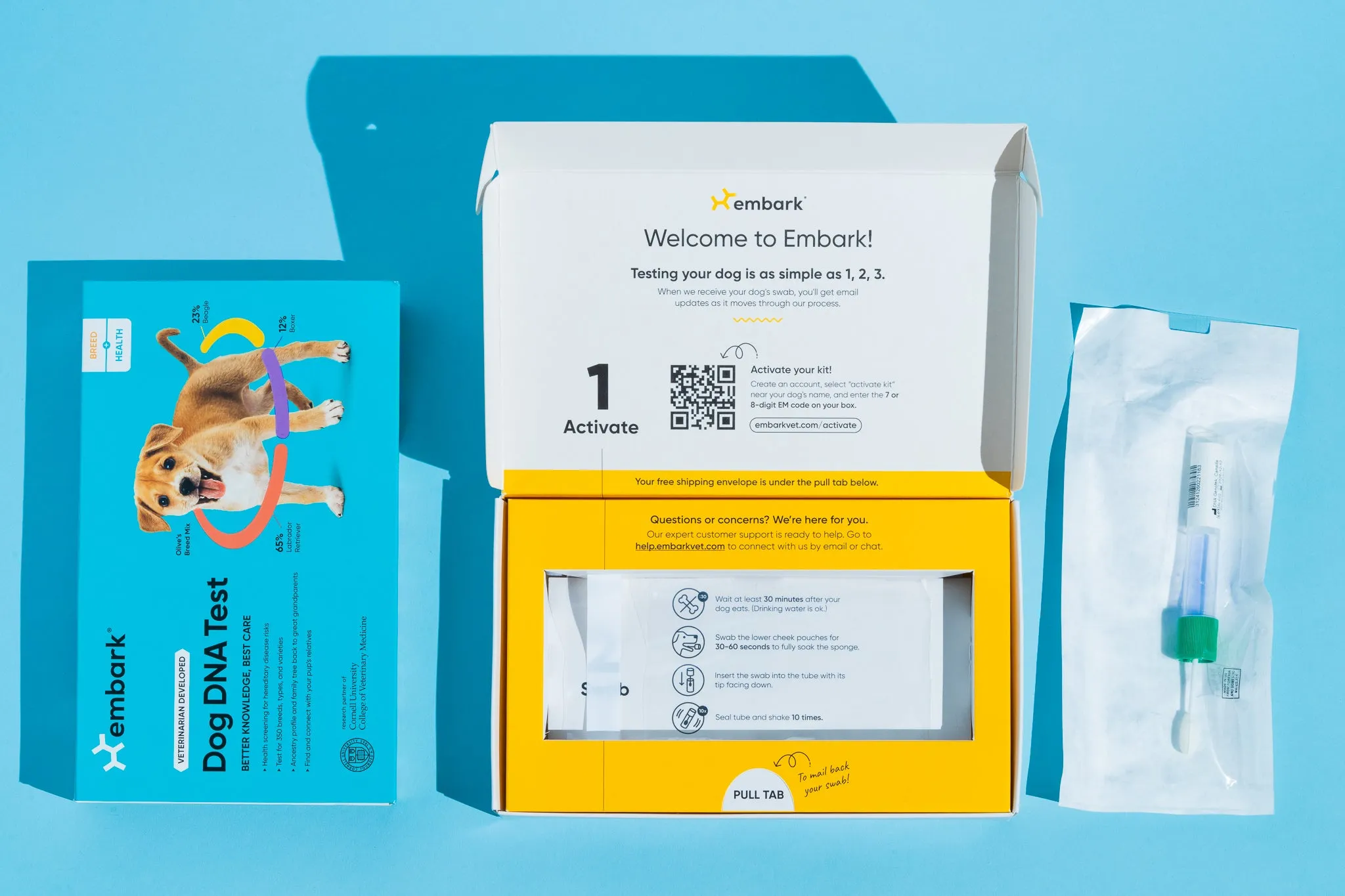 An Embark Dog DNA Test kit displayed showing the blue box, the opened yellow test kit with instructions, and a sealed swab