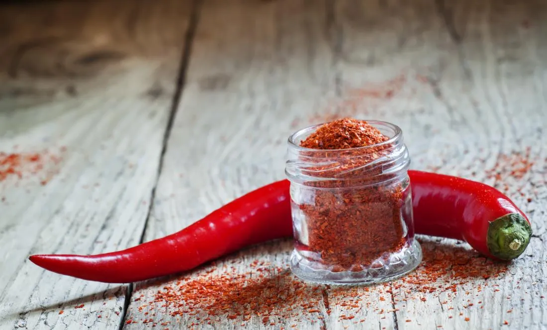 Cayenne pepper behind pot of chili powder on wooden table.