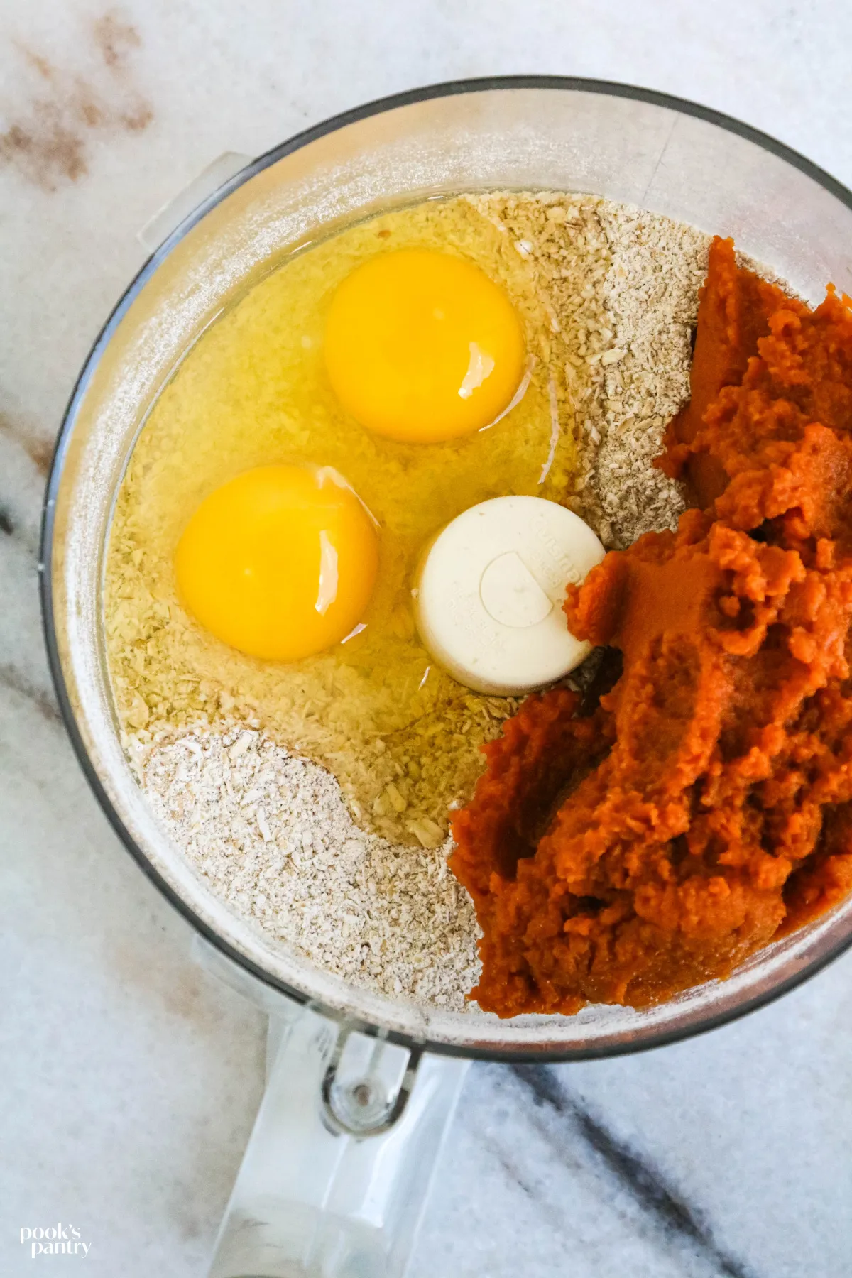 Close-up of old-fashioned oats, pure pumpkin puree, and eggs being combined in a food processor bowl to make dog treat dough.