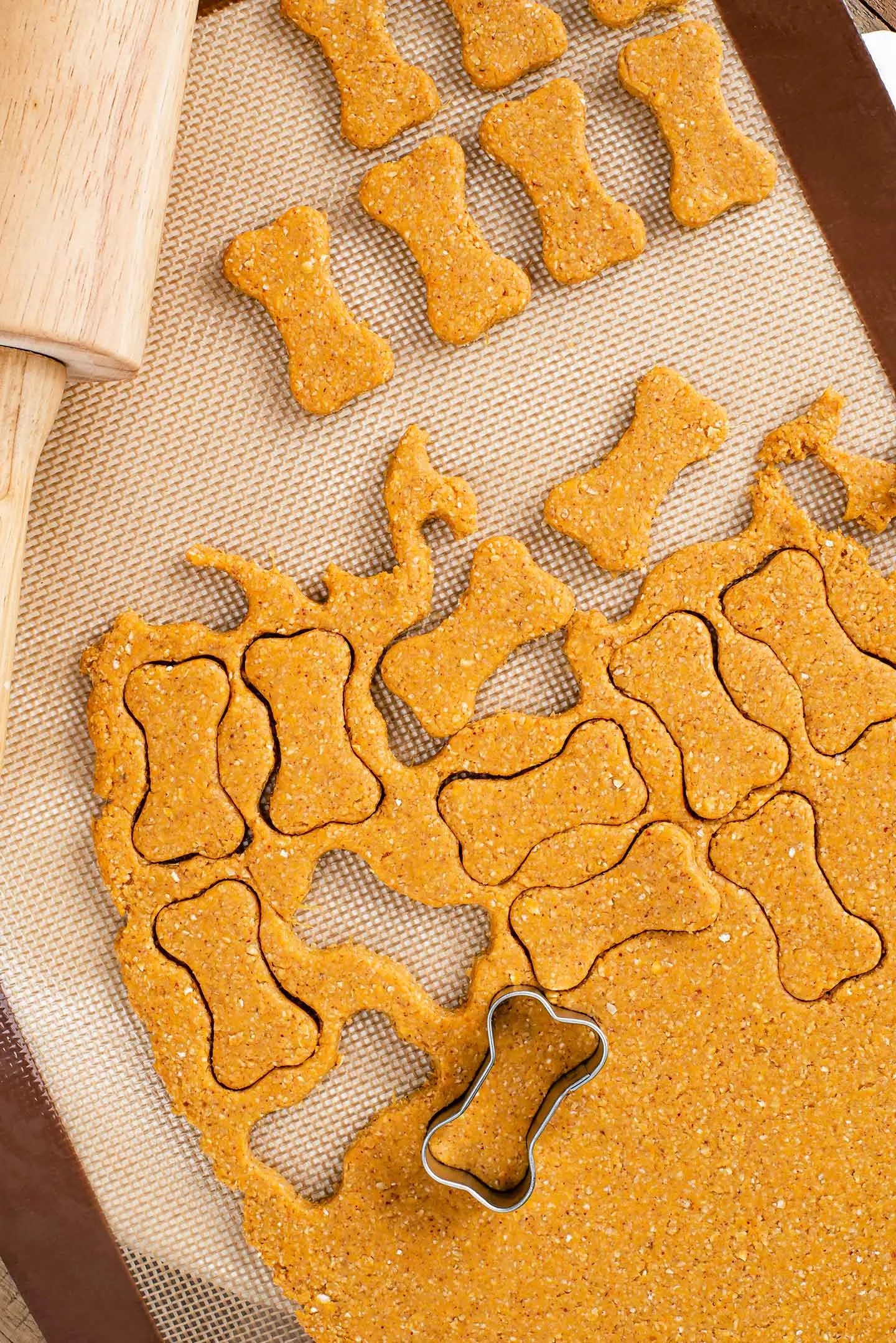 Dog treat dough rolled out on a silicone mat with a rolling pin and bone-shaped cookie cutters.