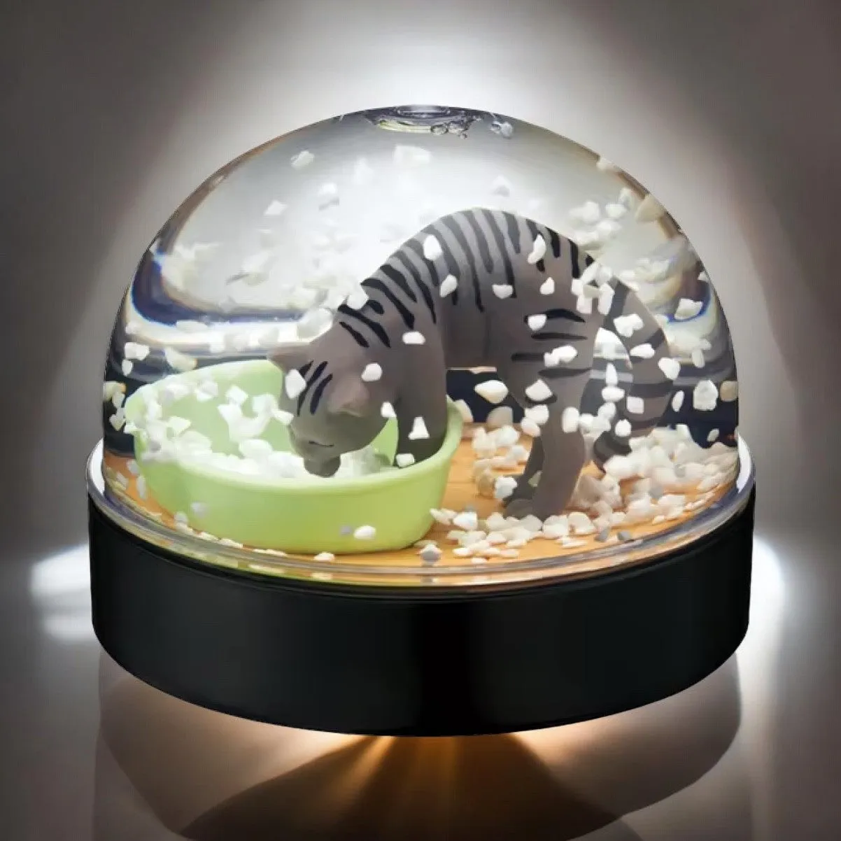 Grey cat playfully making a mess inside a clear snow globe designed as a litter box