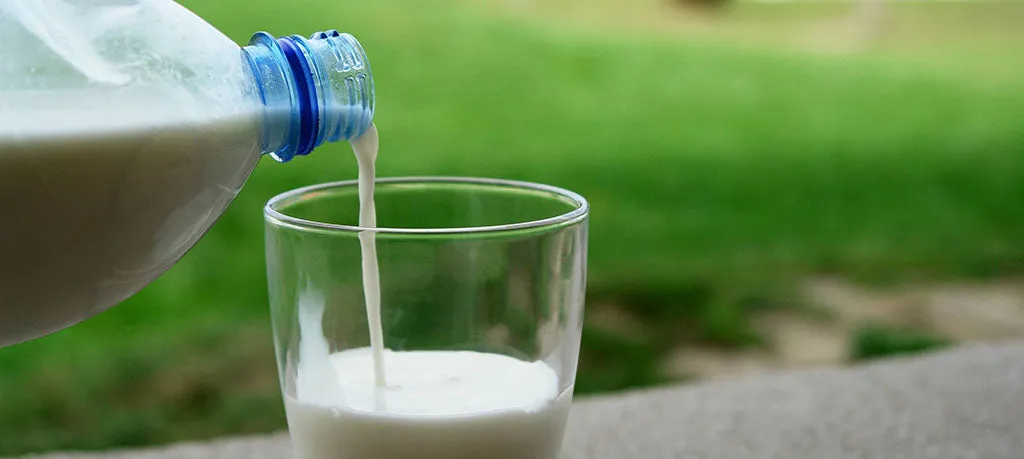 Kefir being poured from a bottle into a clear glass, set against a blurred green outdoor background