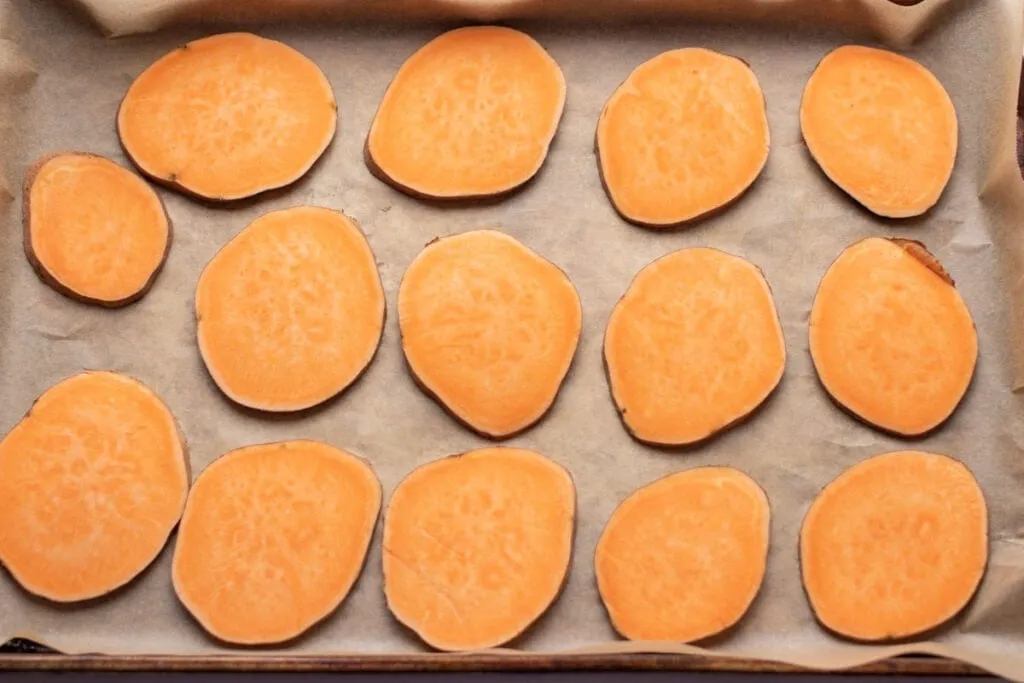 Neatly sliced sweet potato rounds laid out on a baking tray lined with parchment paper, ready for the oven.