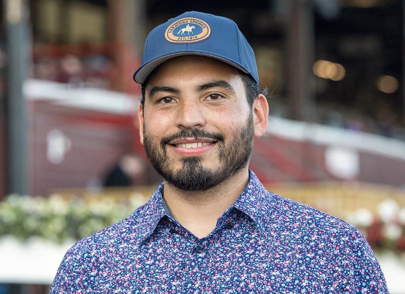 Young, determined horse trainer Jorge Delgado observing his horses during morning workouts, with the racetrack in the background.