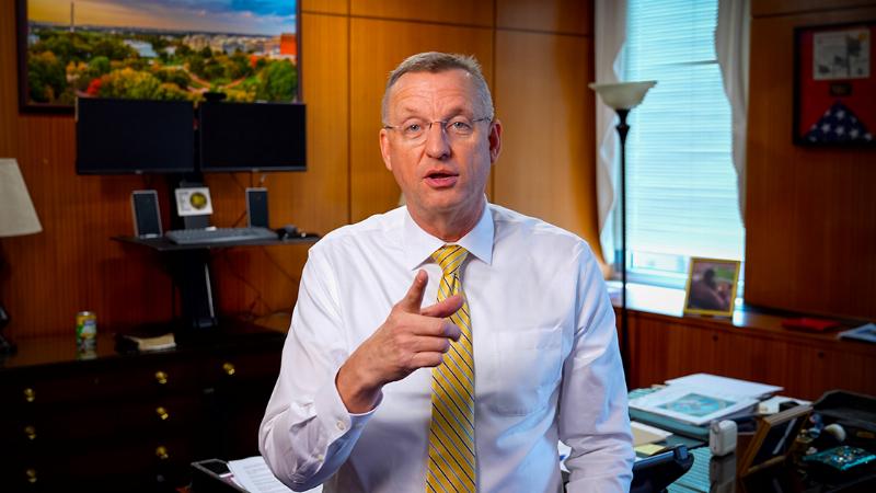A professionally trained service dog, a Golden Retriever wearing a red service vest, sitting calmly beside its handler on a bench with the U.S. Capitol Building visible in the background, illustrating public access in DC.